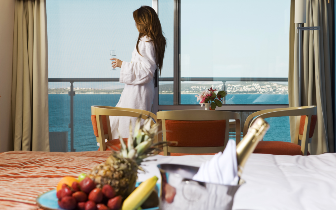 Luxurious hotel room with ocean view, a woman enjoying a drink by the window, and a table set with gourmet food.